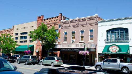 A street view of a downtown area featuring brick buildings, shops, and parked cars under a clear blue sky.