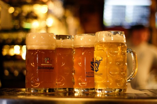 Four glasses of beer with frothy tops, displayed on a bar counter with a warm, blurred background.