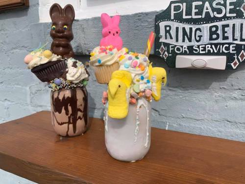 Two colorful milkshakes topped with cupcakes, candy, and marshmallow bunnies, displayed on a wooden shelf.