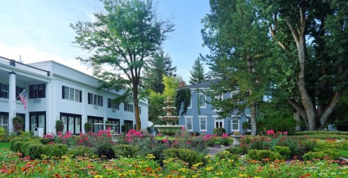 A landscaped garden with colorful flowers and a fountain, surrounded by two elegant buildings and tall trees.