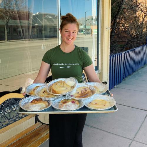 A smiling woman holds a tray of cinnamon rolls outside a bakery, with a sunny street scene in the background.