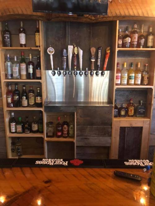 A wooden bar with shelves displaying various bottles of liquor and a row of beer taps on a metal panel.