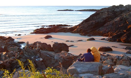 A couple sits on rocky shore, watching the ocean waves at sunset, with stacked stones nearby.