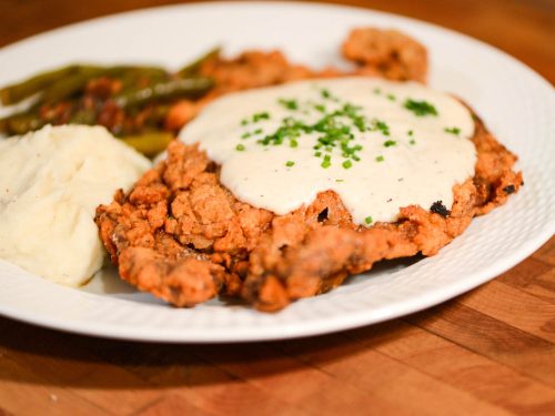 A plate of fried chicken topped with creamy gravy, served with mashed potatoes and green beans.