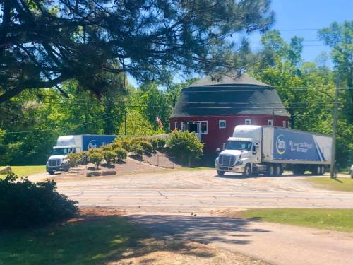 Two large trucks parked near a round, red building surrounded by greenery and trees on a sunny day.