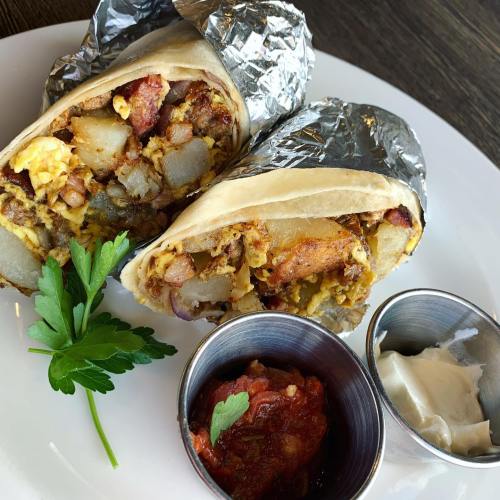 Two breakfast burritos wrapped in foil, served with salsa and sour cream on a white plate, garnished with parsley.