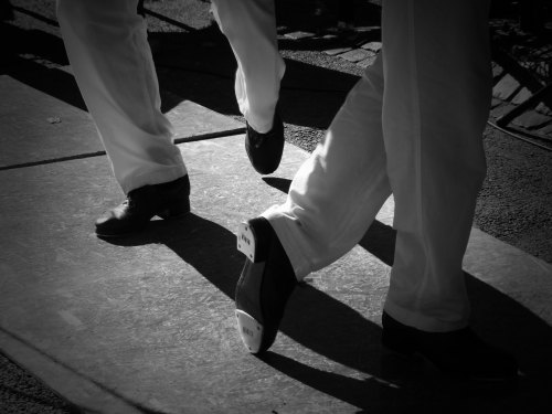 Two pairs of feet in black shoes and white pants walking on a textured surface, captured in black and white.