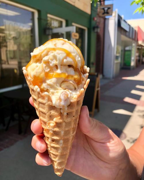 A hand holds a waffle cone filled with creamy ice cream drizzled with caramel, set against a sunny street backdrop.