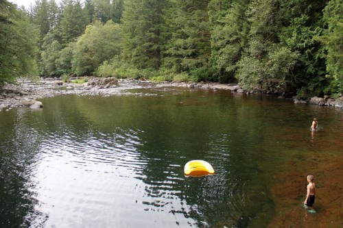 Two children play in a calm river surrounded by lush green trees, with a yellow float nearby.