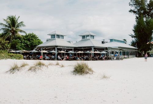 A beachside restaurant with outdoor seating, surrounded by palm trees and sandy beach under a cloudy sky.