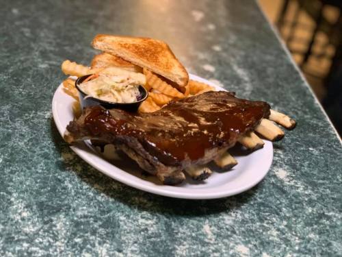 A plate of BBQ ribs with sauce, crinkle fries, coleslaw, and toasted bread on a green speckled table.