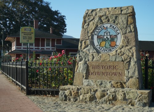 Stone monument with "Historic Downtown" and a colorful emblem, in front of a building and rose bushes in Santa Paula, California.