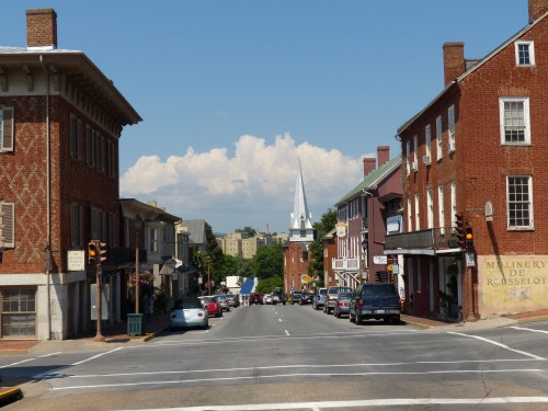 A view of a historic street lined with brick buildings, leading to a church steeple against a blue sky.