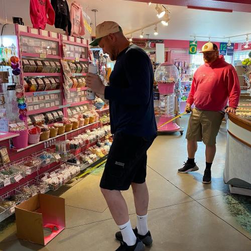 Two men in a colorful candy store, one holding a drink and the other observing, surrounded by shelves of sweets.