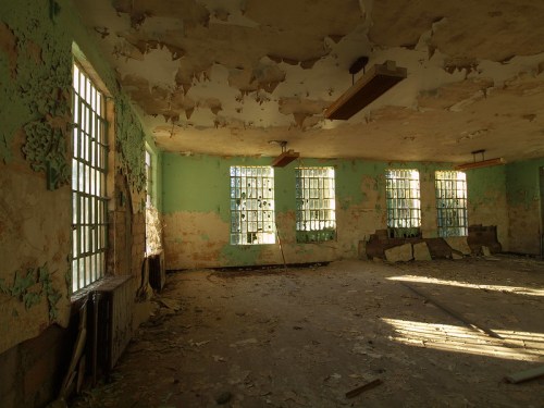 Abandoned room with peeling paint, large windows, and scattered debris, illuminated by natural light.