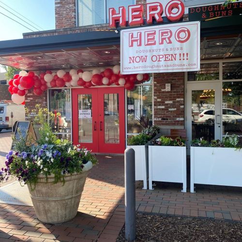 Exterior of Hero Doughnuts & Buns with a "Now Open" sign and colorful balloons above the entrance. Flower planters in view.