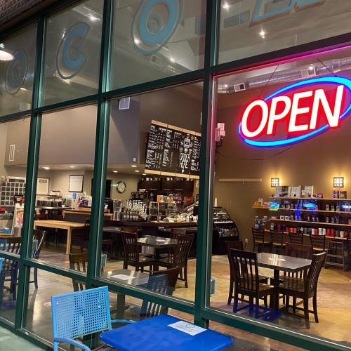 A cozy café with an "OPEN" sign, featuring tables, a menu board, and a coffee counter visible through large windows.