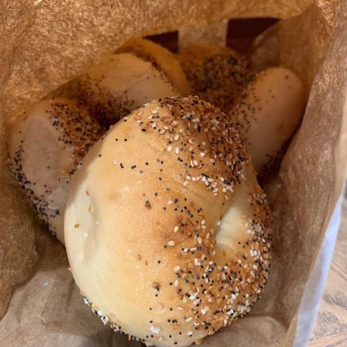 A close-up of several freshly baked bagels with a variety of seeds, placed in a brown paper bag.