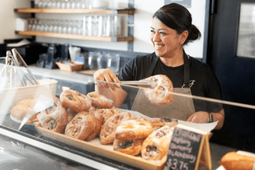 A smiling woman serves pastries from a display case in a bakery, using tongs to pick up a pastry.