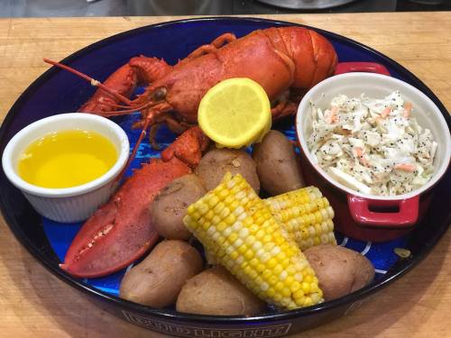 A vibrant seafood platter featuring a lobster, corn, potatoes, coleslaw, and lemon, served with olive oil.