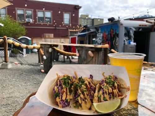 Three tacos topped with slaw and sauce, served with a lime, alongside a glass of beer in a vibrant outdoor setting.