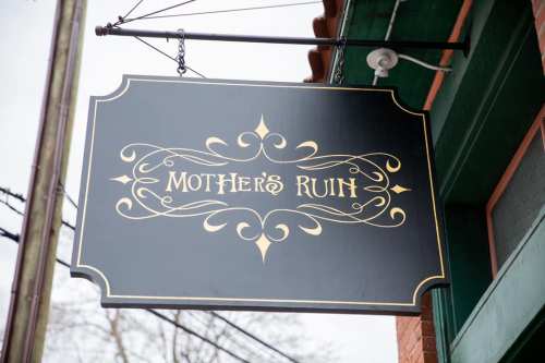 Sign for "Mother's Ruin" featuring elegant gold lettering on a black background, hanging from a building.
