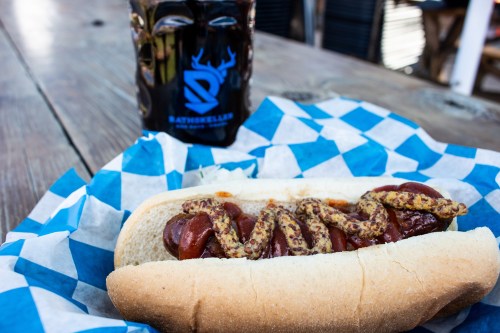 A hot dog in a bun topped with mustard, served on blue and white checkered paper, with a drink in the background.