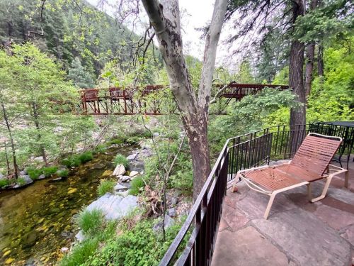 A wooden bridge spans a stream surrounded by lush greenery, with a lounge chair on a patio nearby.