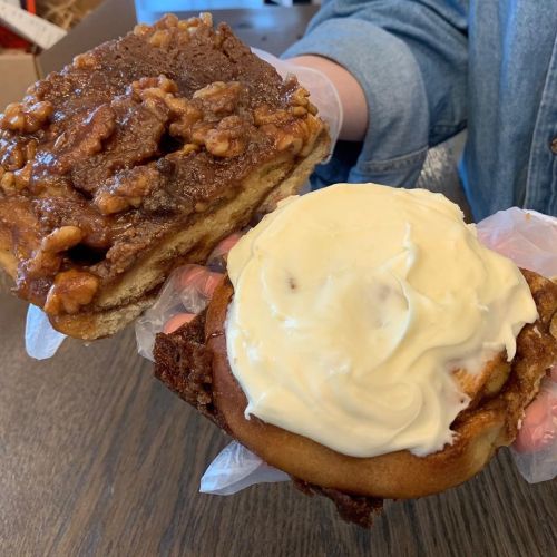 A person holds two cinnamon rolls: one topped with caramel and nuts, the other with cream cheese frosting.