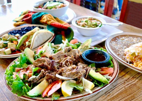 A colorful spread of Mexican dishes, including tacos, salad, and bowls of soup, served on a wooden table.