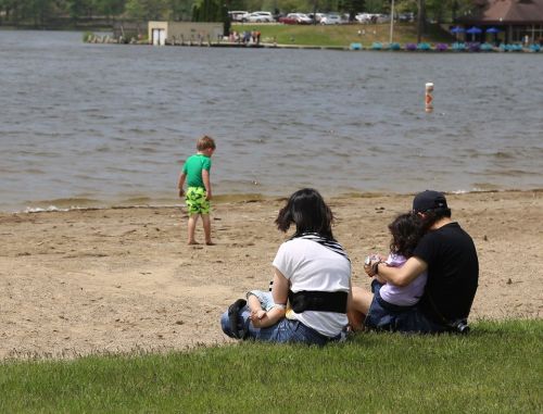 A family sits on the beach while a child plays near the water, with trees and a building in the background.