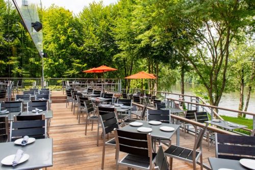 Outdoor restaurant patio with black tables and chairs, orange umbrellas, surrounded by lush greenery and a river view.