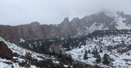 Snow-covered mountains and cliffs surround a winding river in a misty landscape. Pine trees dot the snowy ground.