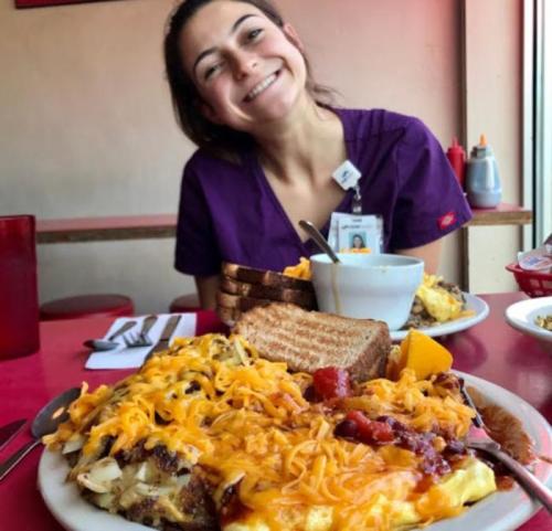 A smiling woman in purple scrubs sits at a table with a large plate of cheesy breakfast food and toast.