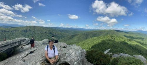 A person sits on a rocky outcrop, overlooking a lush green valley and mountains under a blue sky with fluffy clouds.