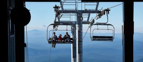 Three people sitting on a ski lift, with mountains and a clear blue sky in the background.
