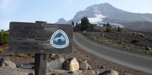 Sign for the Pacific Crest National Scenic Trail with a mountain backdrop and a winding road leading into the distance.