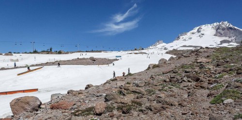 A snowy mountain landscape with skiers and snowboarders, rocky terrain, and a clear blue sky in the background.