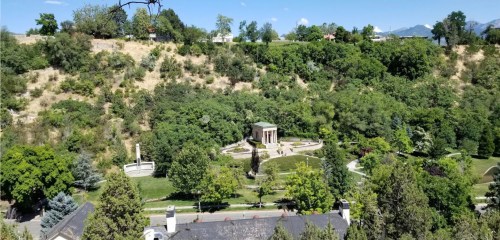 Aerial view of a green park featuring a circular monument surrounded by trees and pathways. Mountains in the background.