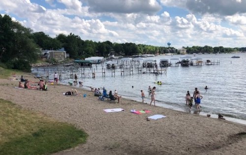 A sandy beach with people relaxing, swimming, and boats docked in the water under a partly cloudy sky.