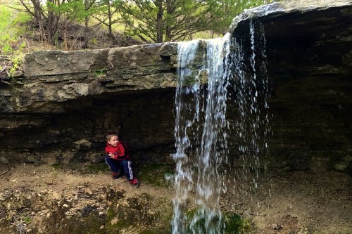 A young boy sits beside a small waterfall cascading over rocky terrain in a natural setting.