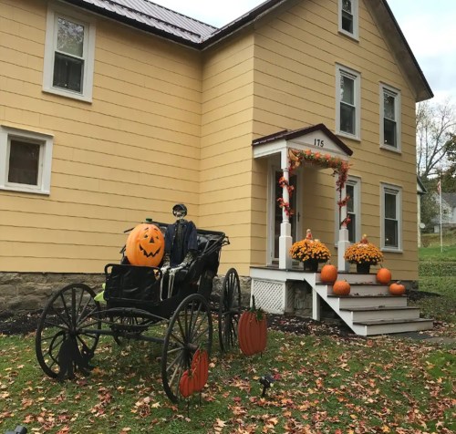 A yellow house decorated for Halloween, featuring pumpkins, a skeleton in a carriage, and autumn flowers.