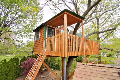 A wooden treehouse with a balcony, perched in a tree, surrounded by greenery and a sloped roof below.