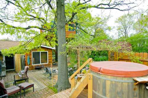 A backyard scene featuring a hot tub, wooden deck, patio chairs, and a treehouse among lush greenery.