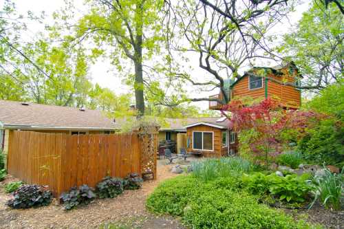 A cozy backyard with a wooden fence, lush greenery, and a treehouse among trees and flowering plants.