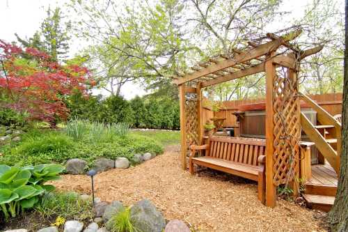 A wooden pergola with a bench in a lush garden, surrounded by greenery and colorful plants, with a hot tub nearby.