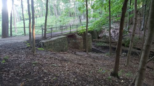 A stone bridge over a small creek, surrounded by trees and a forested area with a path nearby.