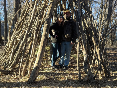 A couple stands together in front of a rustic wooden structure made of branches in a wooded area.
