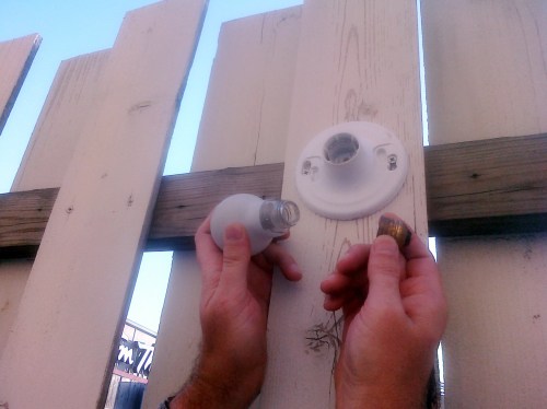 A person installs a light bulb into a fixture mounted on a wooden fence.
