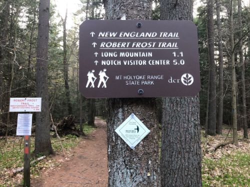 Trail sign in a forest, indicating directions to New England Trail and Robert Frost Trail, with distances to landmarks.